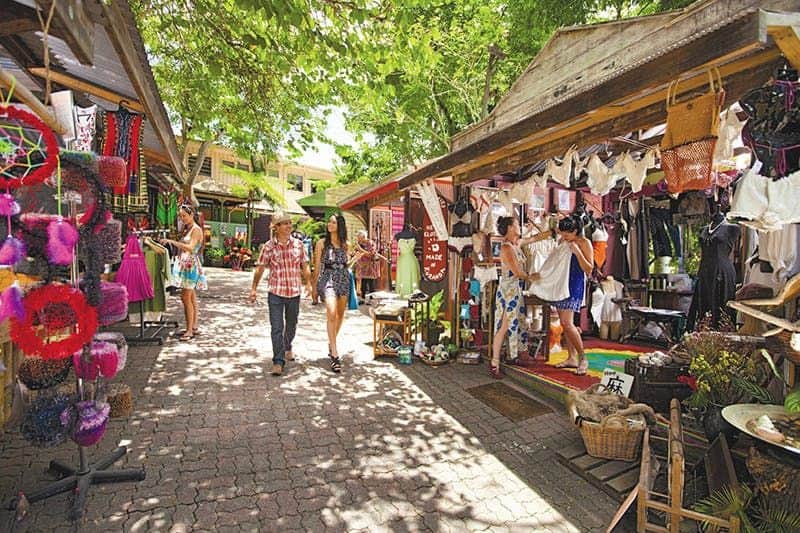 Outdoor market with people shopping for clothing and accessories under shaded stalls surrounded by trees.