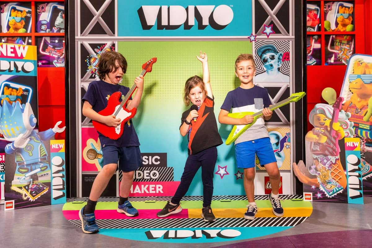 Three children are posing with toy guitars and a microphone in front of a colorful backdrop with the word "VIDIYO."