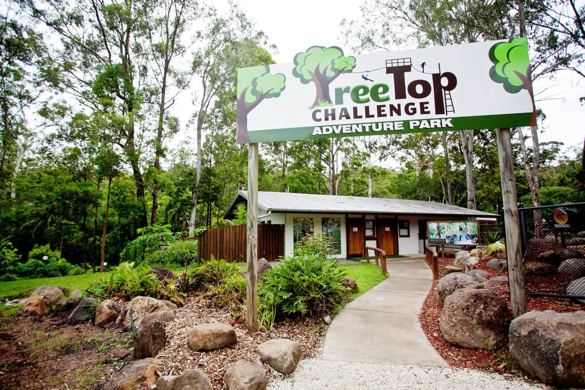 Entrance to TreeTop Challenge Adventure Park with a sign above a pathway leading to a small building surrounded by trees and rocks.