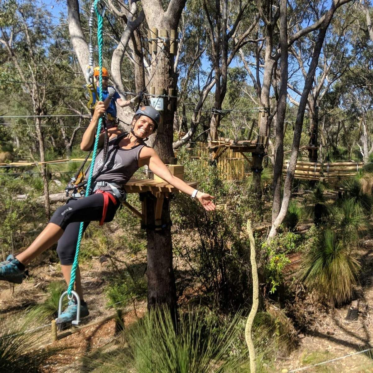 A person smiles and waves while suspended on a rope course in a forested area filled with trees and greenery.