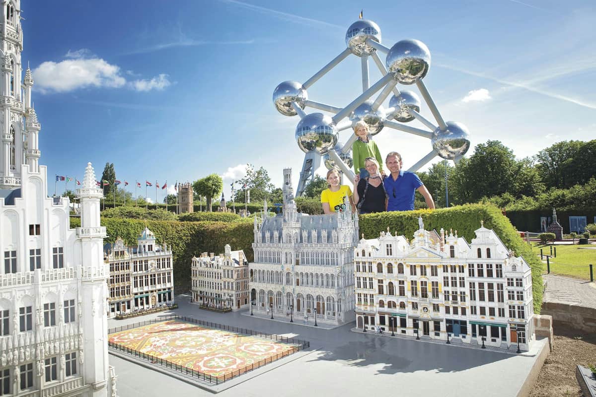 Four people standing behind miniature models of European buildings with the Atomium structure in the background on a sunny day.