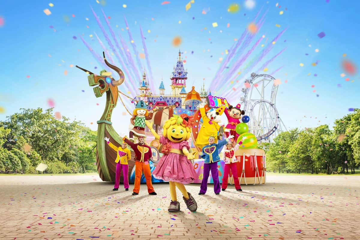 A group of colorful characters in costumes pose in front of a themed castle backdrop, with confetti and a rollercoaster visible.