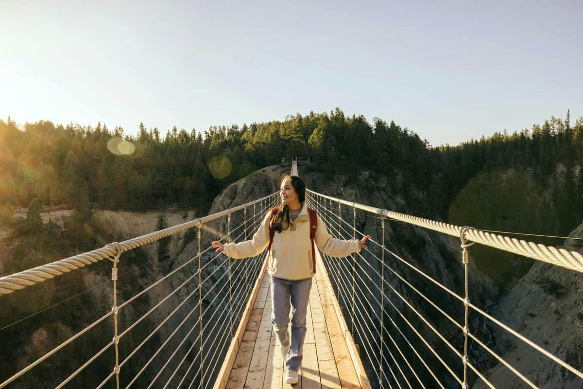 A person walks on a suspension bridge above a forested canyon, holding onto the side ropes and looking to the right.