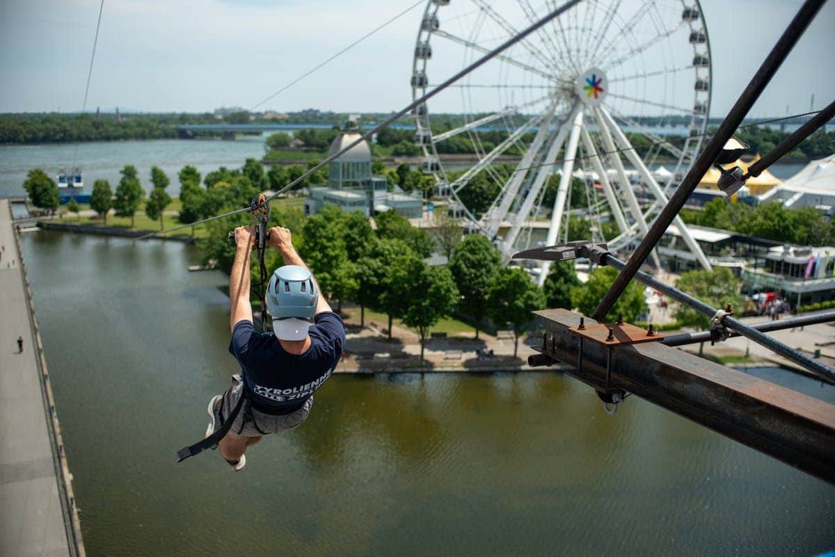 Person ziplining over a body of water with a Ferris wheel and green trees in the background.