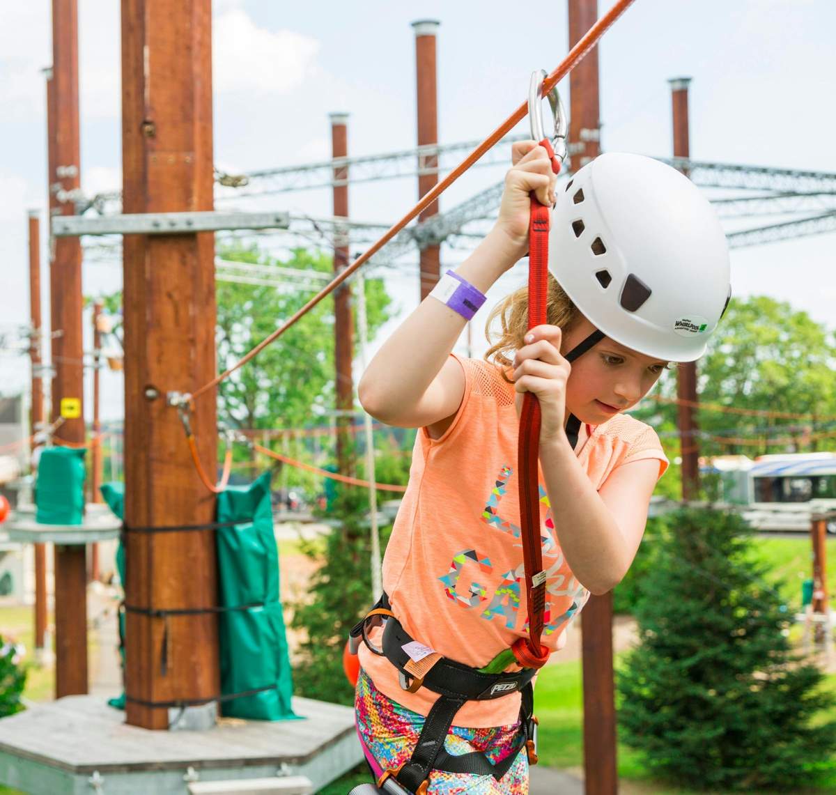 A child wearing a white helmet and harness navigates a ropes course outdoors, holding onto a red safety line.
