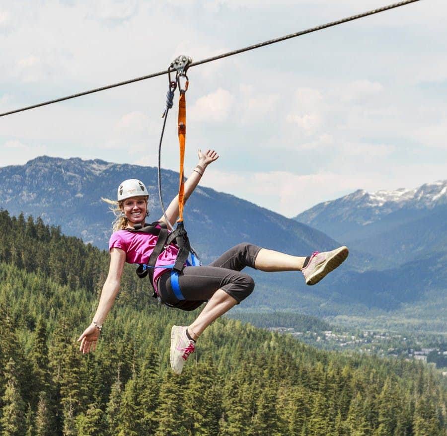A person wearing a helmet and harness is ziplining above a forest with mountains in the background.