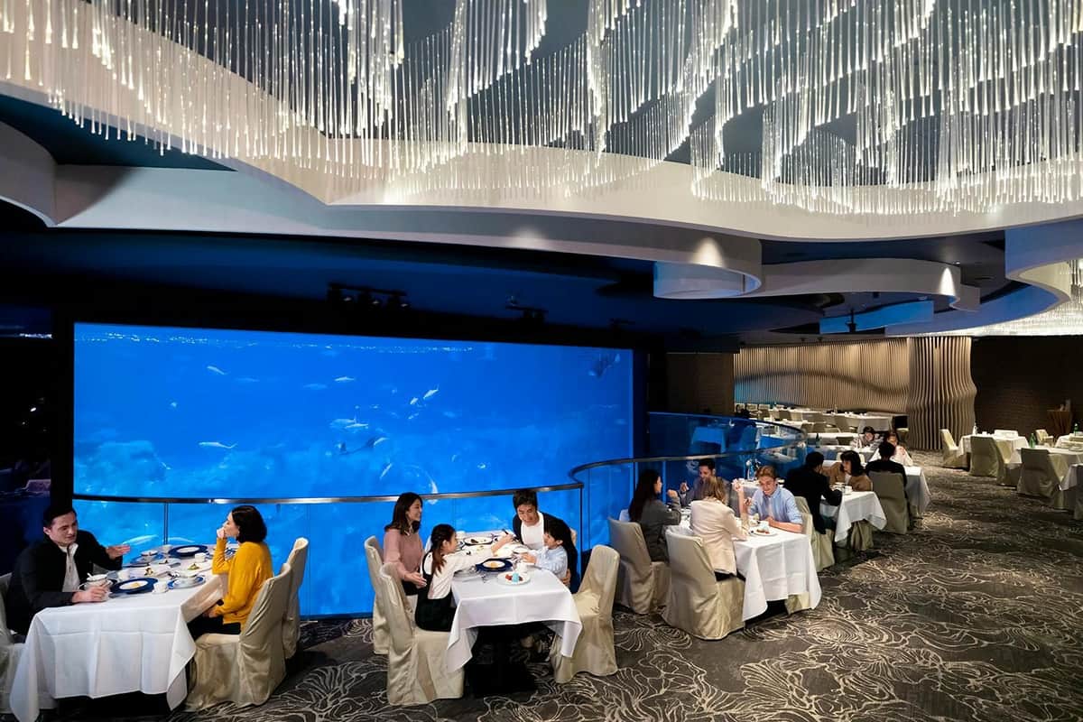 A restaurant with diners seated at tables, featuring a large aquarium wall and a decorative ceiling with hanging lights.