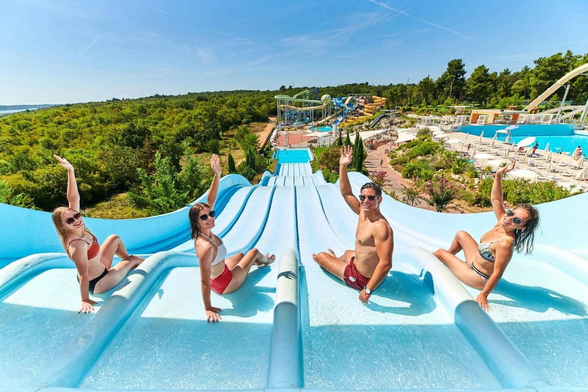 Three people waving at the top of blue water slides with a water park, trees, and blue sky in the background.