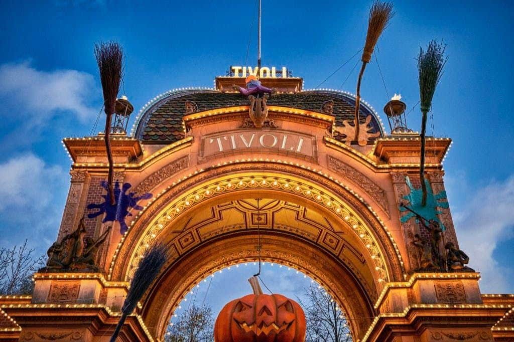 Ornate archway entrance decorated for Halloween with a large pumpkin, witch figure, and brooms, illuminated by lights.