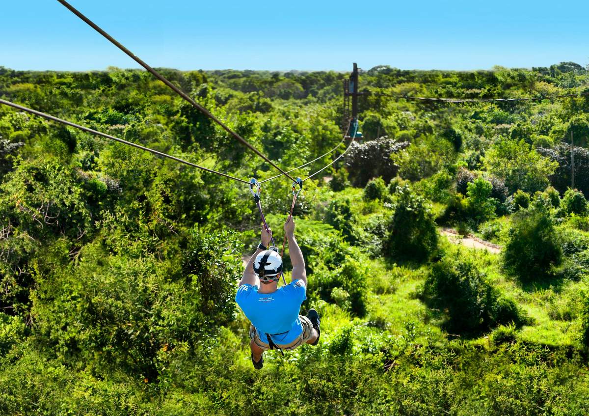 Bavaro Park: Buggy + Zipline Person ziplining over lush green forest, wearing a blue shirt, shorts, and helmet. Sky is clear with bright sunlight.