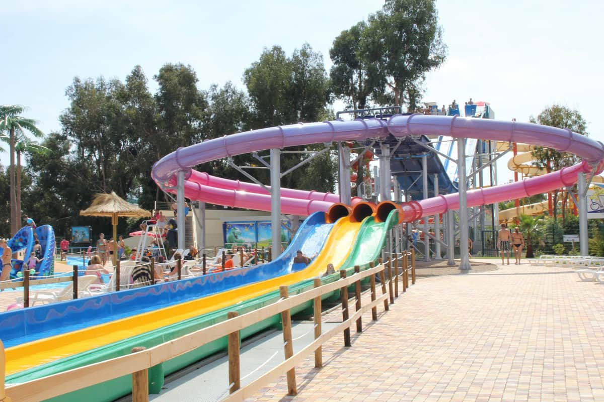 Aquasplash: Entry Ticket Colorful water slides in an outdoor water park with people in the background, surrounded by trees and a paved walkway.