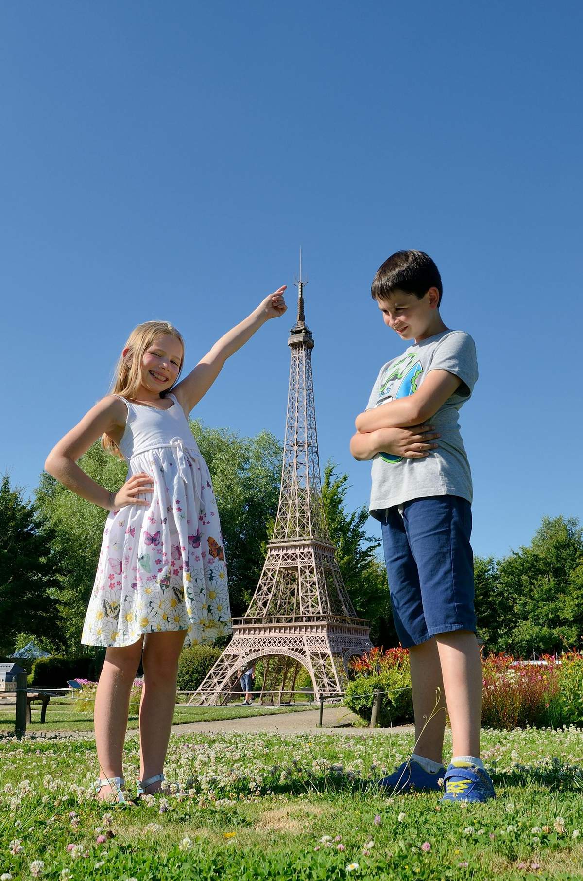 A girl points and smiles at a miniature Eiffel Tower while a boy stands beside her with arms crossed, both outdoors on a sunny day.