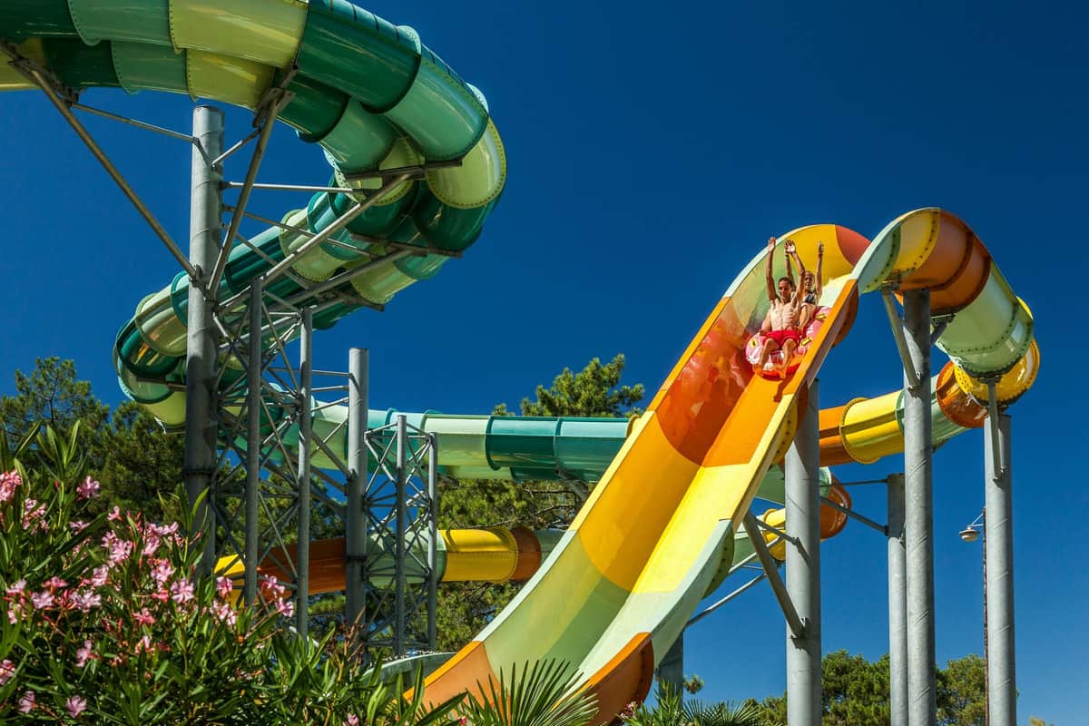 Aqualand Arcachon: Entrada Dos personas bajando por un colorido tobogán acuático en un parque acuático, bajo un cielo azul claro y rodeadas de vegetación.