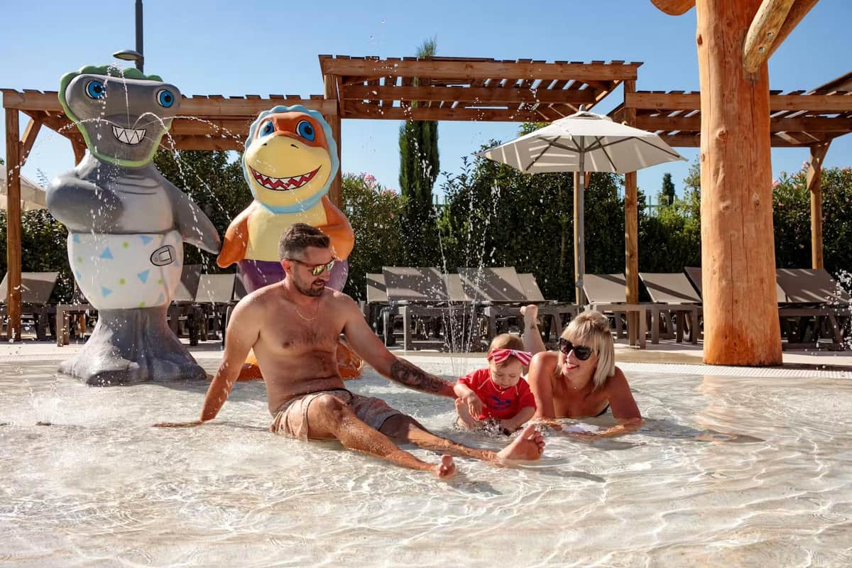 Wave Island: Early Access Ticket – Wave Island (Monteux) Een man, een vrouw en een klein kind spelen in het ondiepe water bij een kleurrijk haaienbeeld en ligstoelen onder parasols.