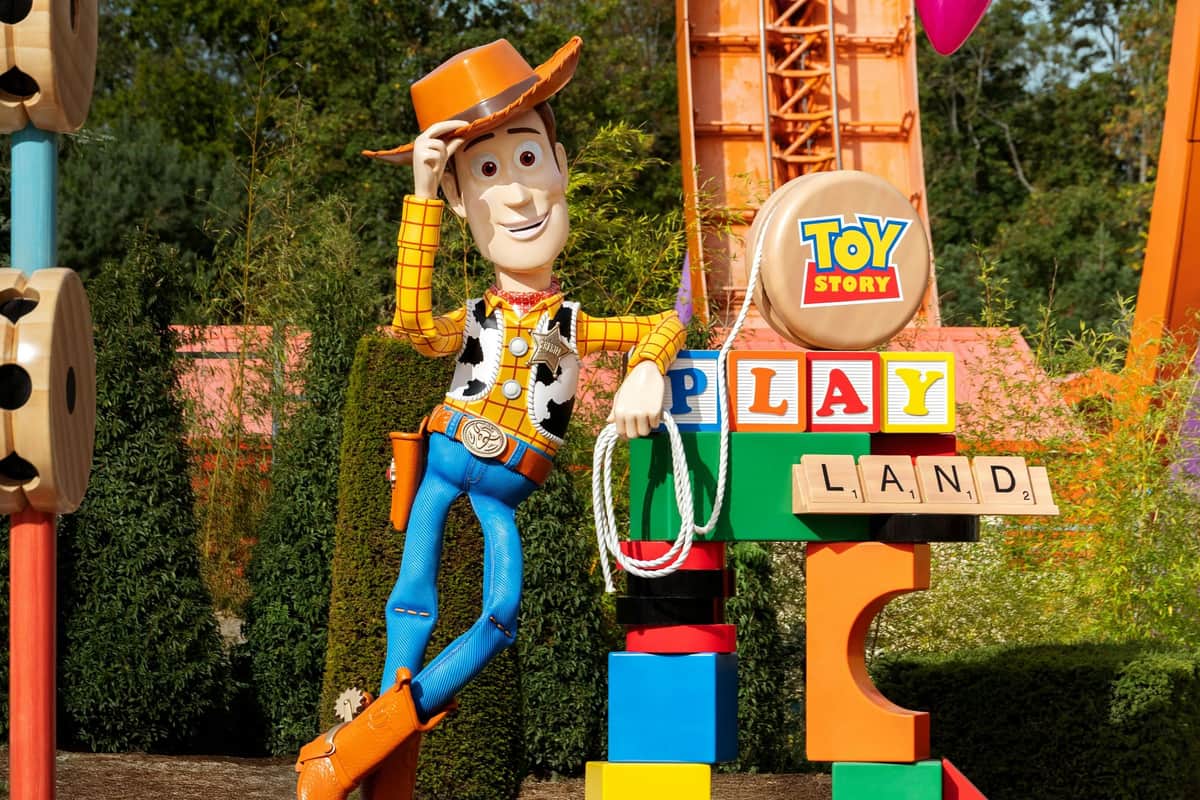 A statue of Woody from Toy Story, standing with one foot raised, next to colorful blocks and greenery in the background.