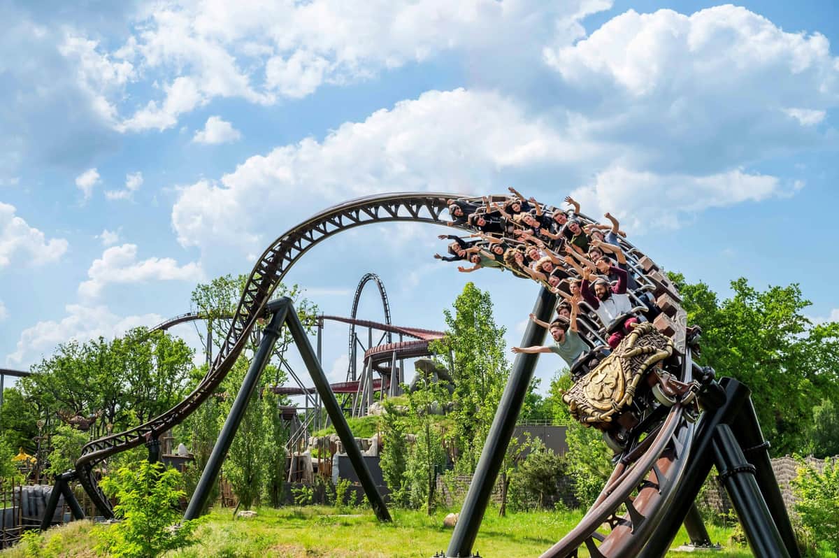 A roller coaster with passengers riding through a loop against a backdrop of greenery and a cloudy sky.