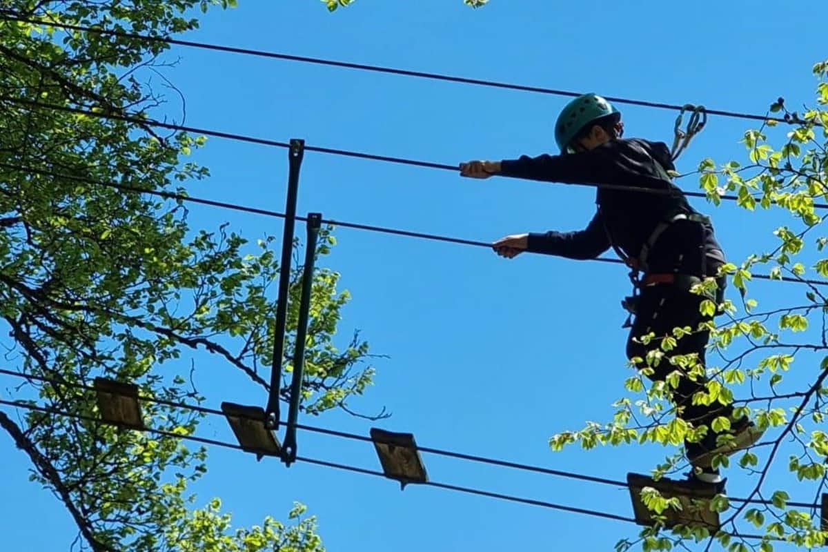 Person in a helmet and harness navigating a high ropes course among trees and a clear blue sky.