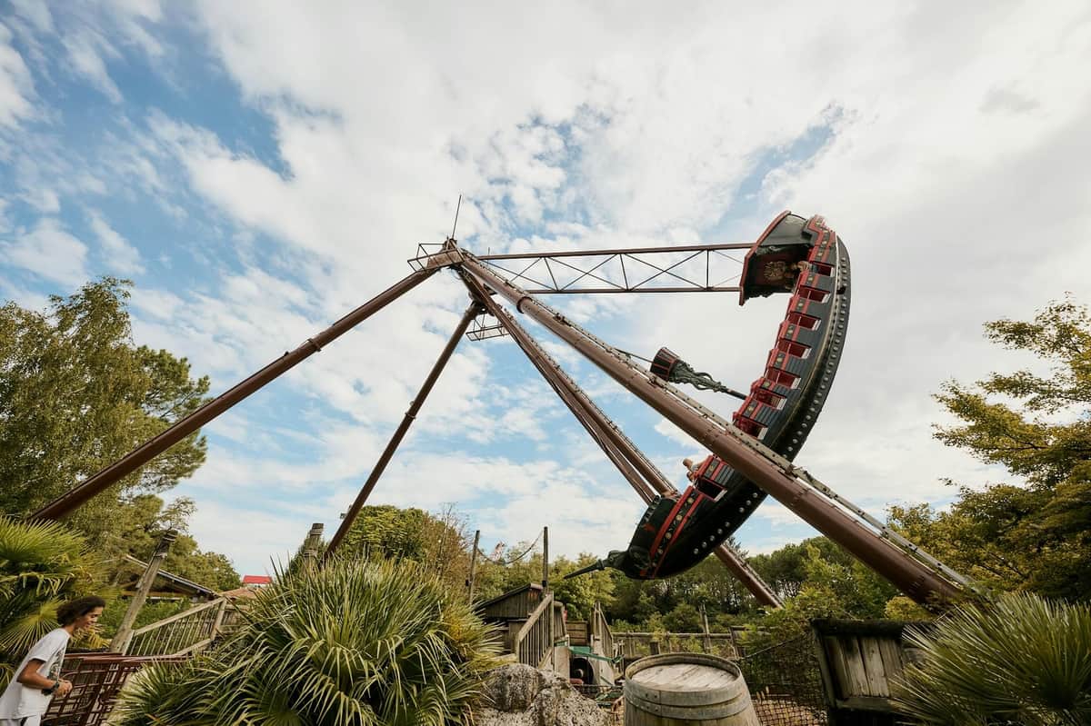 A large pirate ship ride swings high in the air at an amusement park, surrounded by trees and a partly cloudy sky.