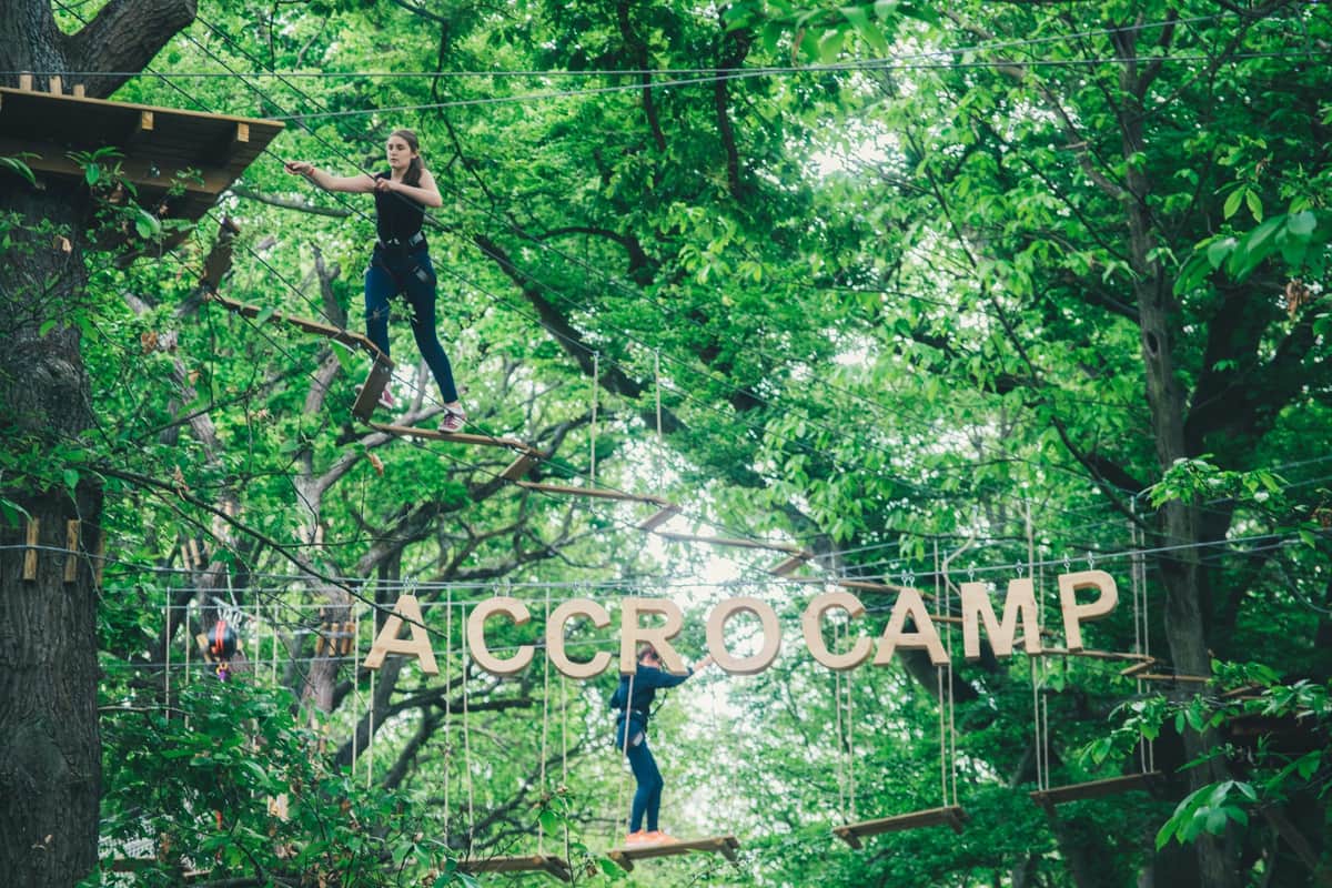 Two people navigate a ropes course amid lush green trees; one stands on a plank, while another balances near wooden letters spelling "ACCRO."
