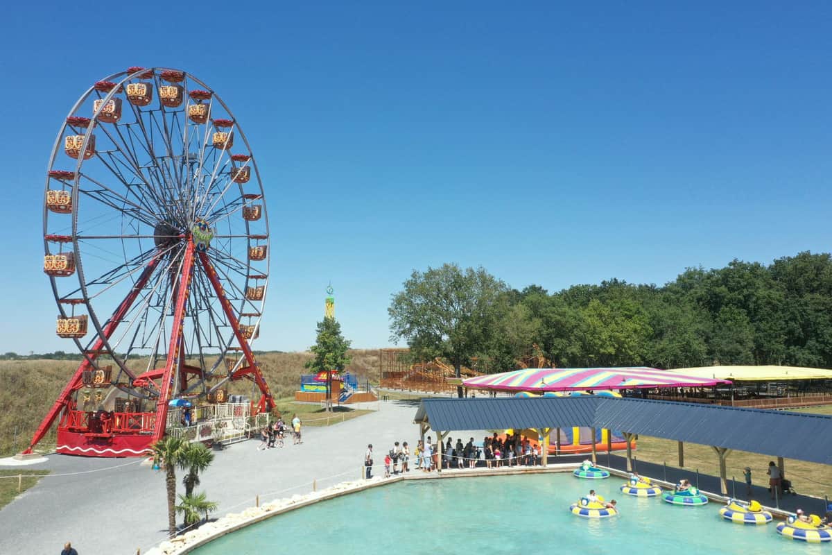Colorful ferris wheel, bumper boats in a pool, and a striped tent surrounded by trees under a clear blue sky at an amusement park.