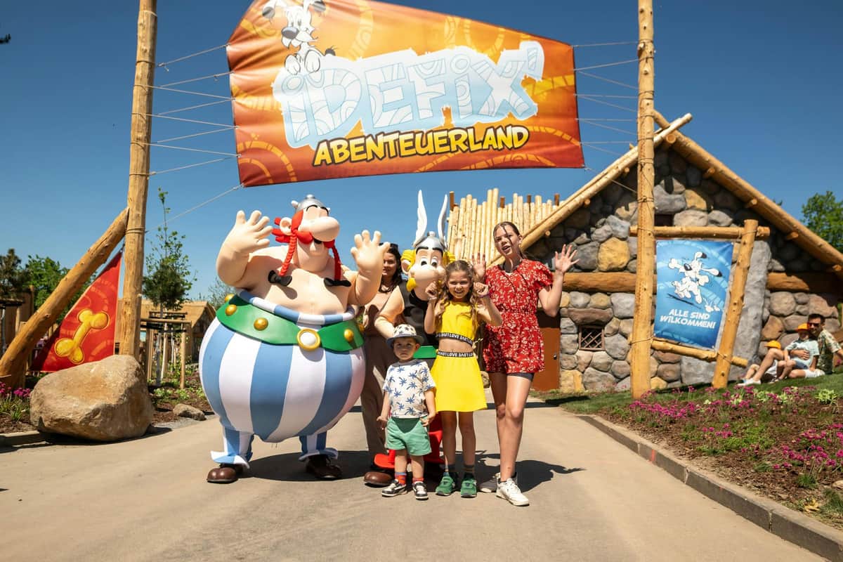 A group of people posing with costumed characters in front of a sign reading 'Idéfix Abenteuerland' and a themed building.