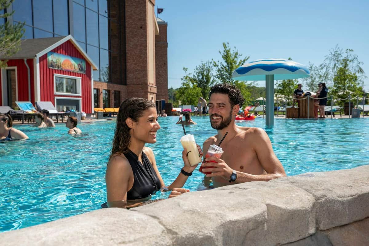 Two people smiling in a swimming pool, holding drinks. Others swim nearby. Red building and umbrella-shaped structure in the background.