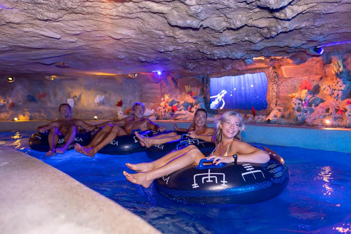 People smiling while floating on blue inner tubes in an indoor aquatic cave with artificial marine decorations.