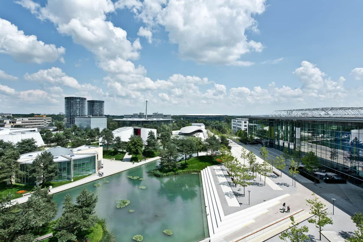 Modern campus with buildings, a pond with lily pads, trees, and a spacious walkway under a partly cloudy sky.