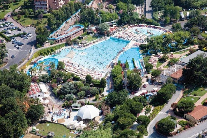 Aerial view of a bustling water park with several pools, water slides, umbrellas, loungers, and lush greenery.