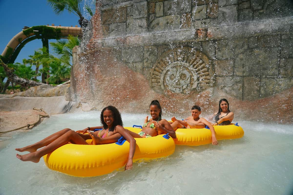 Four people in yellow inflatable tubes float under a waterfall with a stone wall featuring a decorative carving in the background.