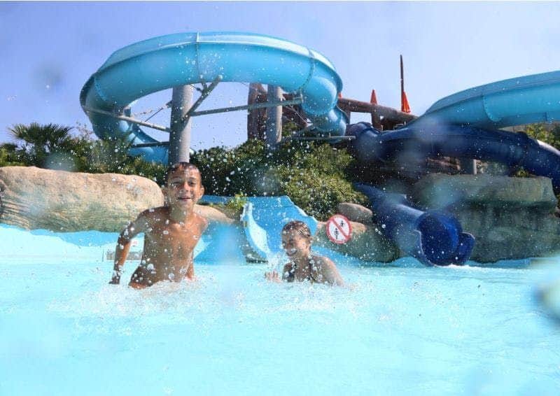 Two boys playing and splashing in a pool below a large blue waterslide on a sunny day.