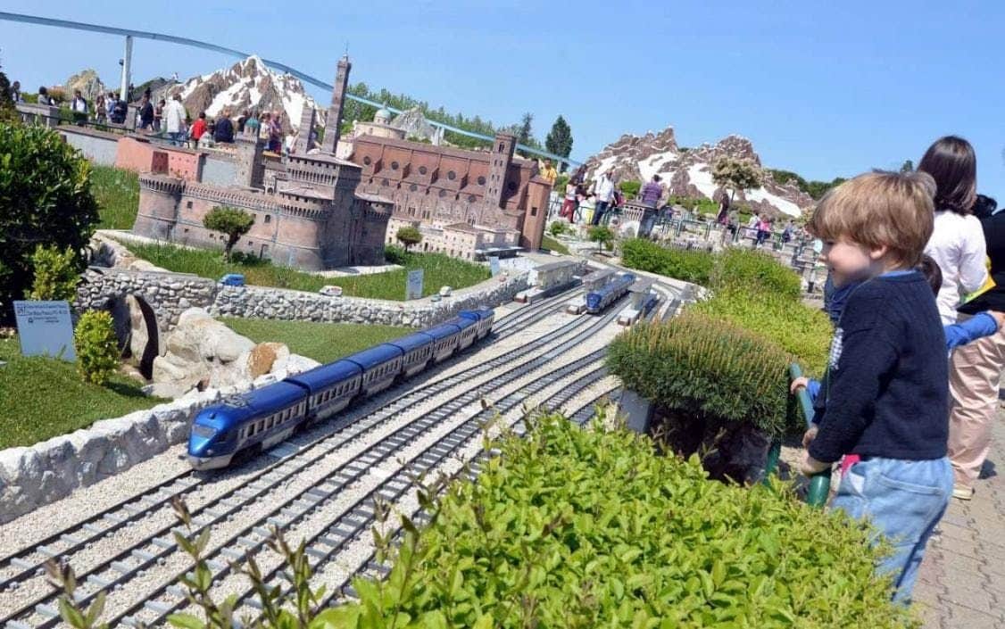 People observing a large outdoor model train set with detailed buildings and landscapes under a clear blue sky.