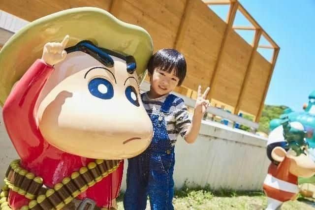 A child posing with a large cartoon character statue outdoors near a wooden structure on a sunny day.