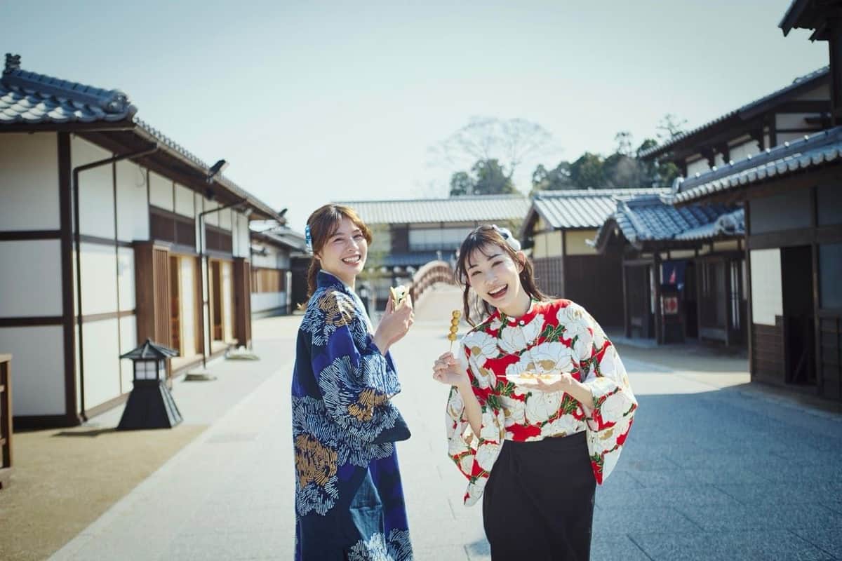 Toei Kyoto Studio Park: Entry Ticket Two women in traditional Japanese attire smile and hold skewers of food, standing on a street with wooden buildings.