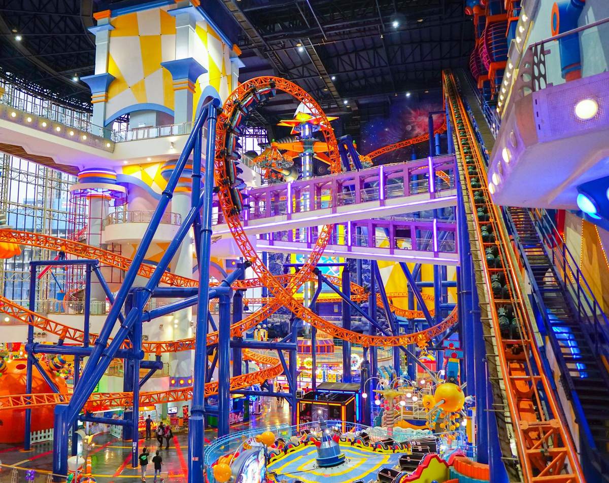 Indoor amusement park featuring a large, colorful rollercoaster with loops and tracks, surrounded by other rides and attractions.