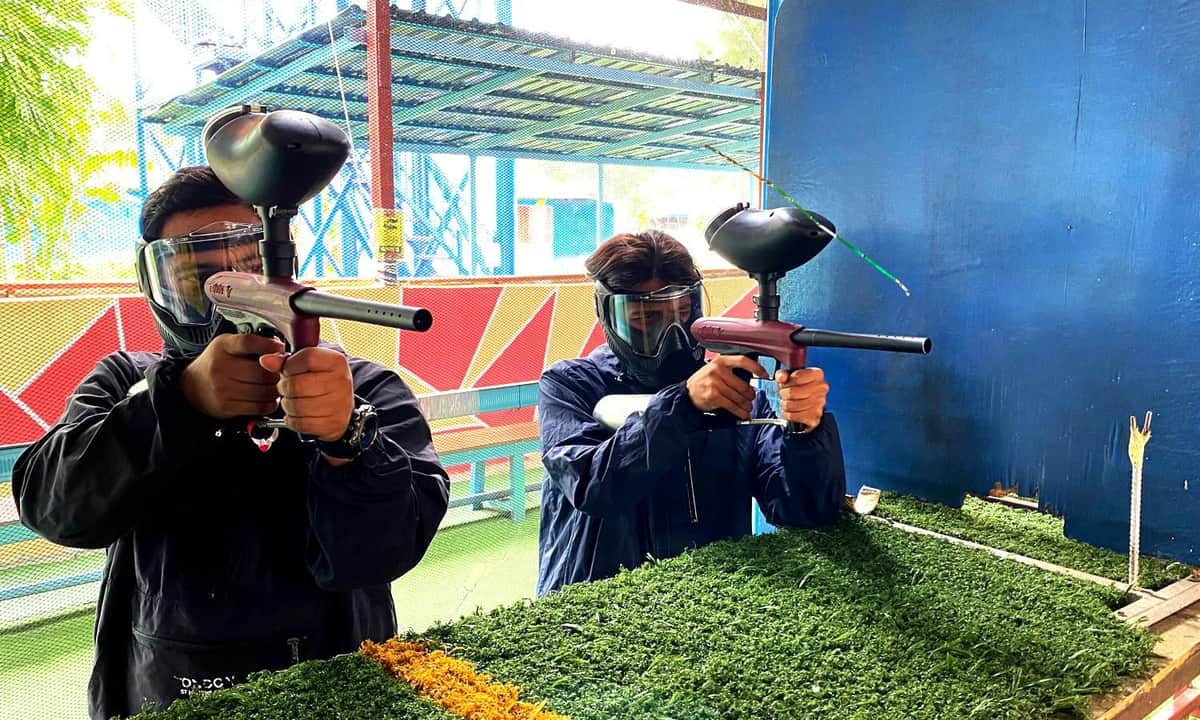 Two individuals wearing protective masks and holding paintball guns, standing in an outdoor area with colorful surroundings.