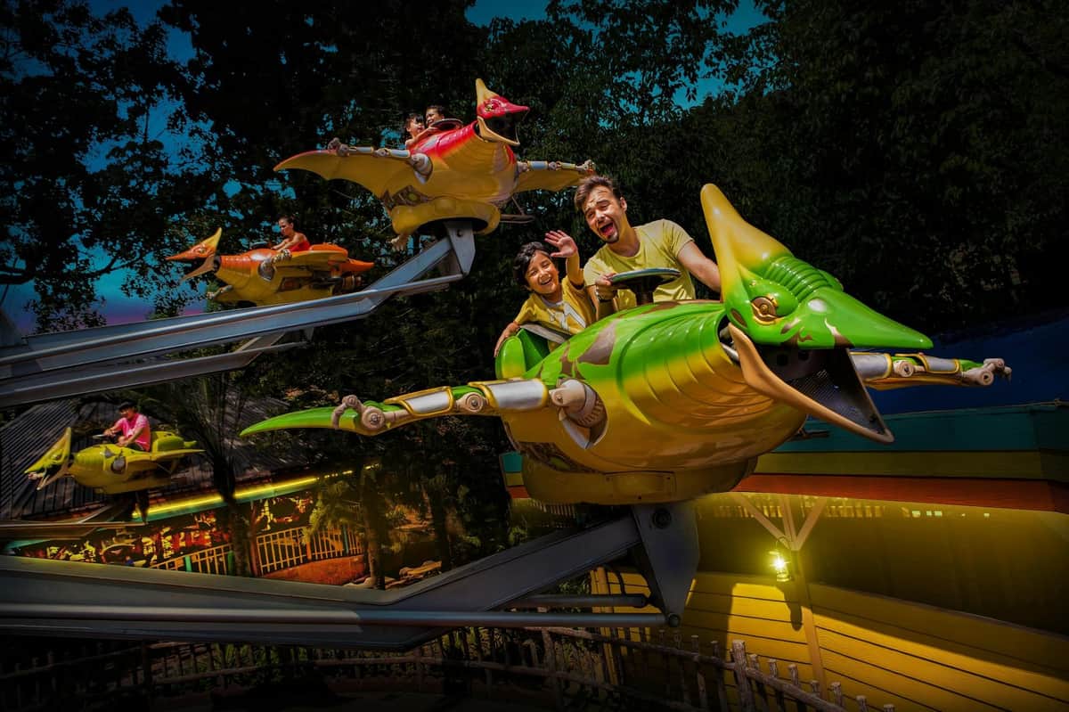 Sunway Lagoon: Night Entry Ticket People enjoying a night-time amusement park ride on pterodactyl-themed vehicles, with trees and lit buildings in the background.