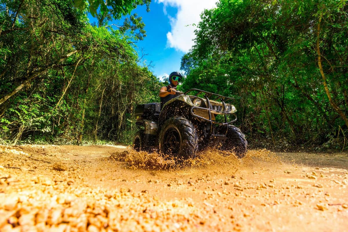 Person riding an ATV through a muddy trail surrounded by lush greenery under a clear blue sky.