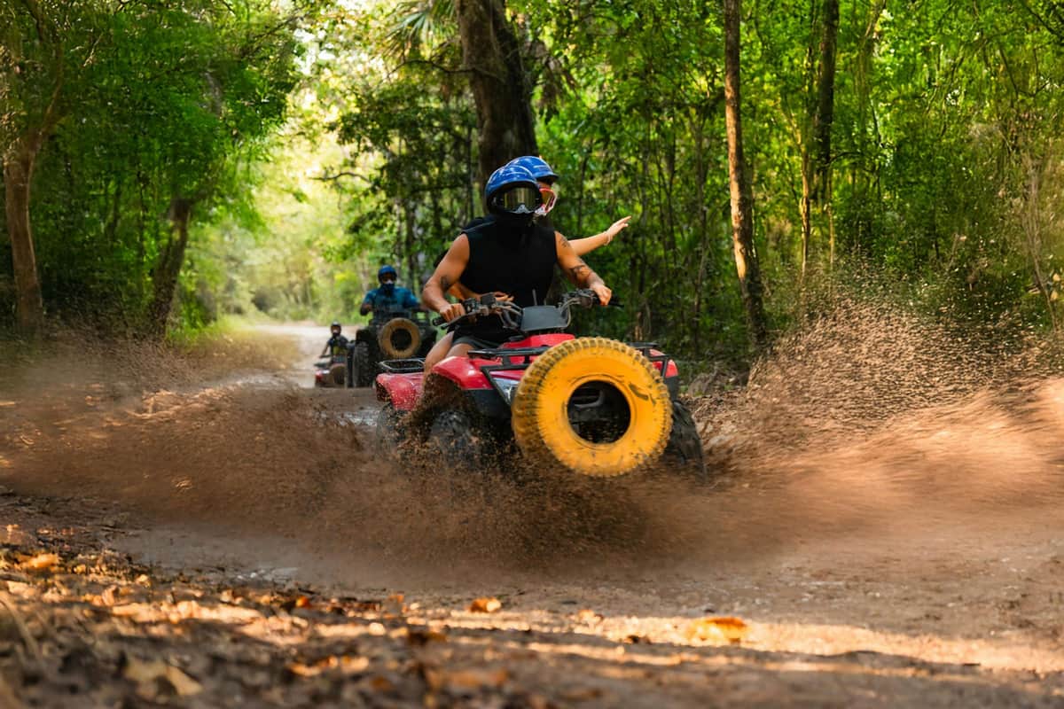 Mayan Jungle Rush: ATV + Ziplines + Lunch + Transfer A person wearing a helmet and bandana rides an ATV on a dirt path, surrounded by trees and greenery.