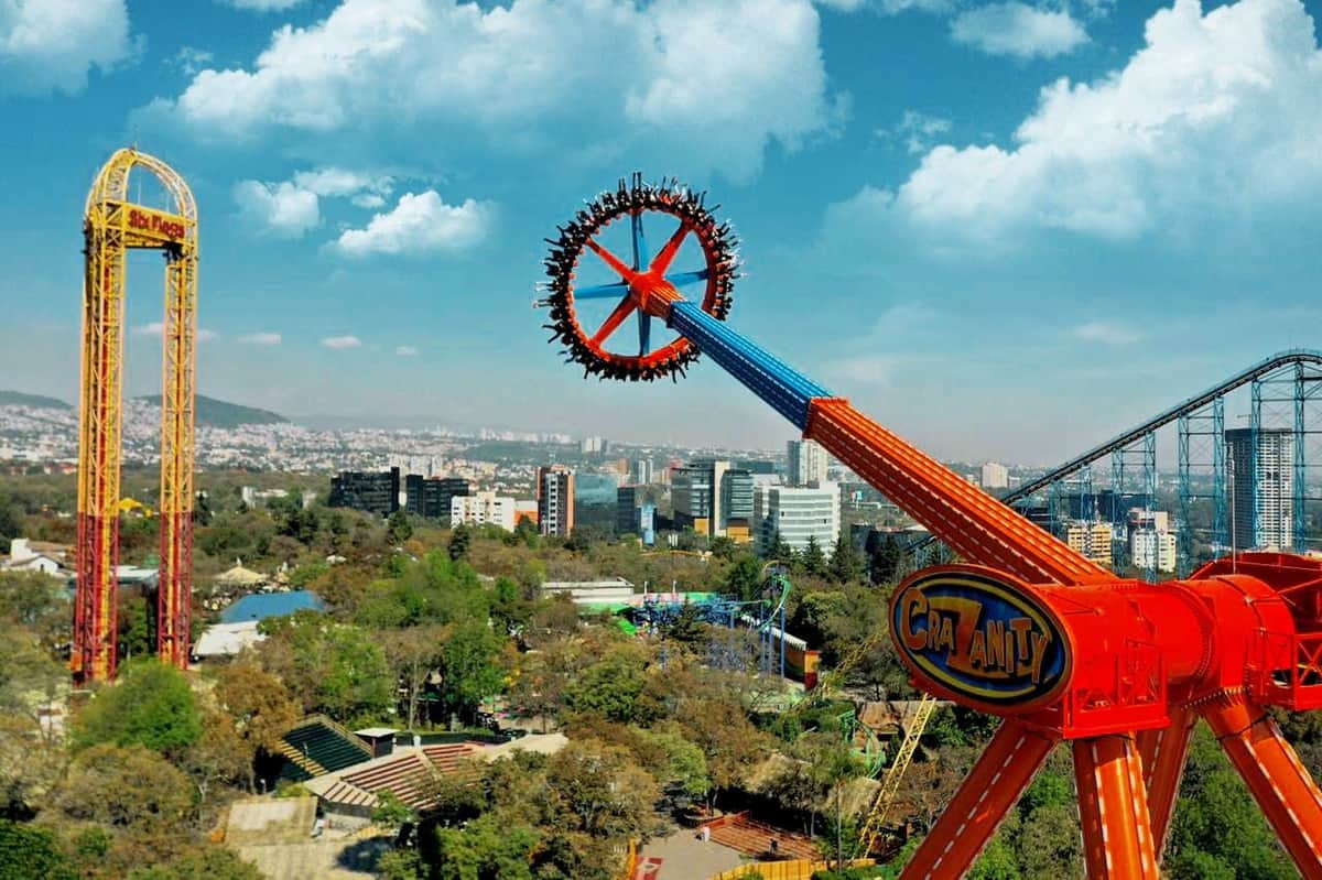 Six Flags Mexico: Day Pass Wesołe miasteczko jedzie w ruchu, wirując wysoko nad pejzażem miejskim z budynkami, drzewami i błękitnym niebem z chmurami w tle.