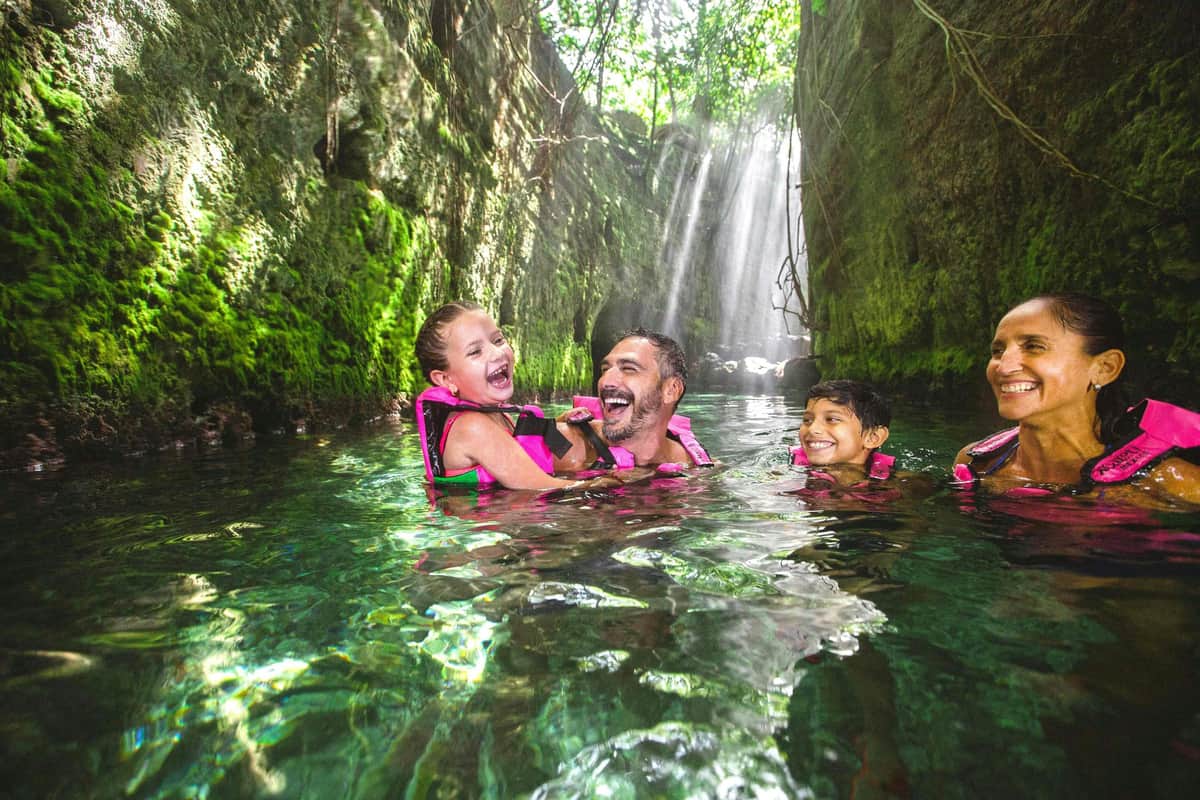 Xcaret Park: Entry Ticket A family of four wearing life jackets smiles and enjoys swimming in a green canyon with sunlight streaming through trees above.