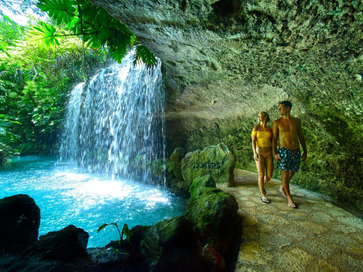 Xenses Park: Entry Ticket A couple walks on a stone path near a waterfall inside a cave, with lush greenery visible overhead.