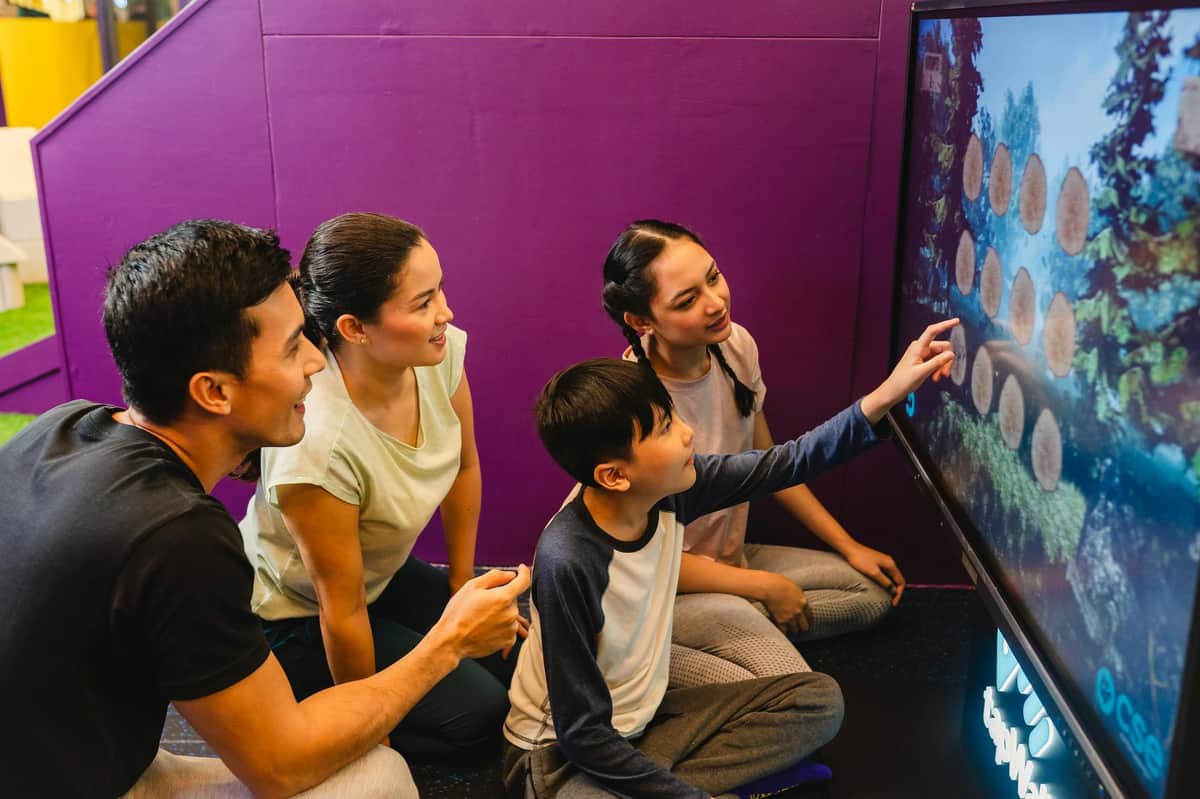 A group of four people, two adults and two children, sitting on the floor and smiling, interacting together indoors.