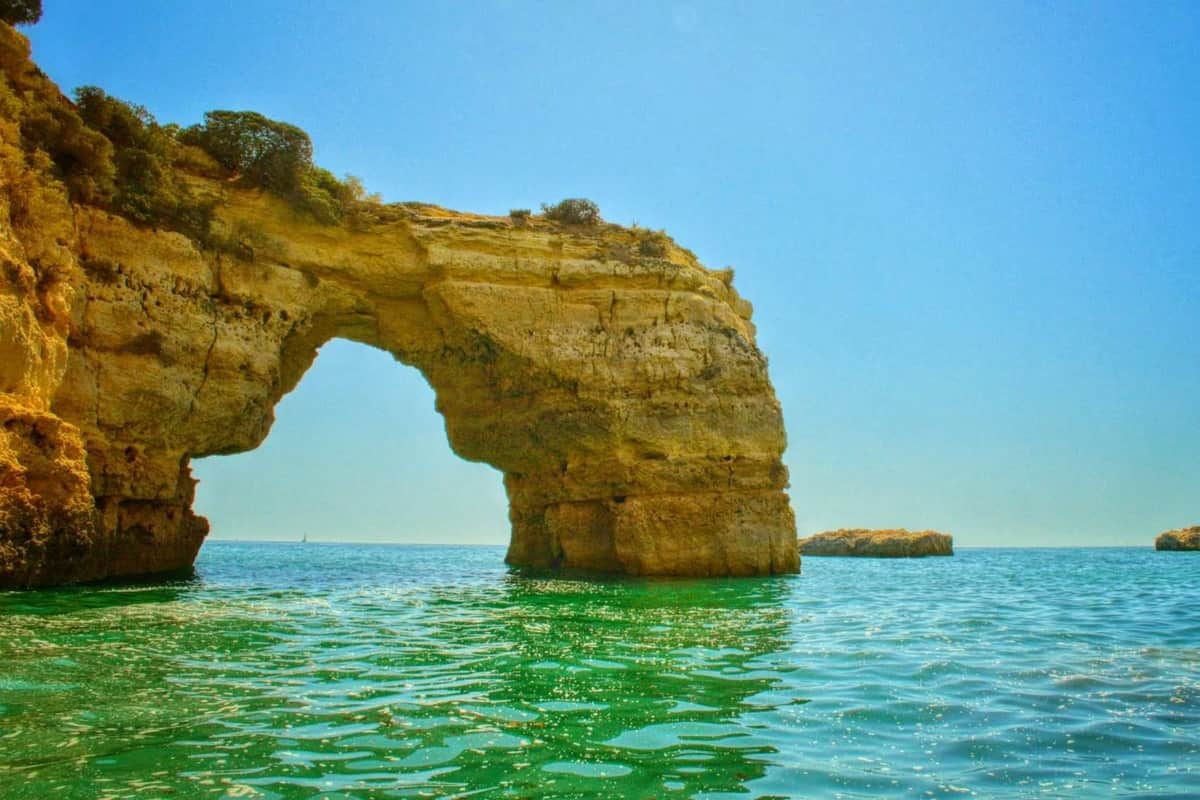 Rock formation with a natural arch rising from turquoise seawater under a clear blue sky. A smaller rock is visible in the background.