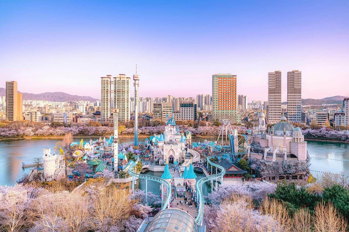 Lotte World: Entry Ticket Amusement park with colorful rides and attractions, surrounded by cherry blossoms, with tall city buildings in the background.