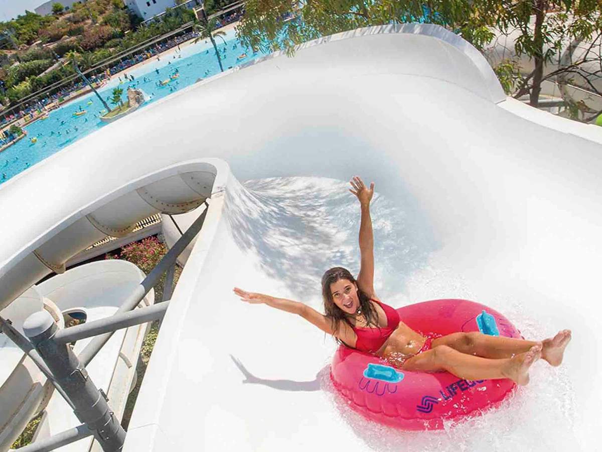 Aqua Natura Benidorm: Entry Ticket Woman in a red bikini on a pink inflatable tube sliding down a white water slide, with a pool and trees in the background.