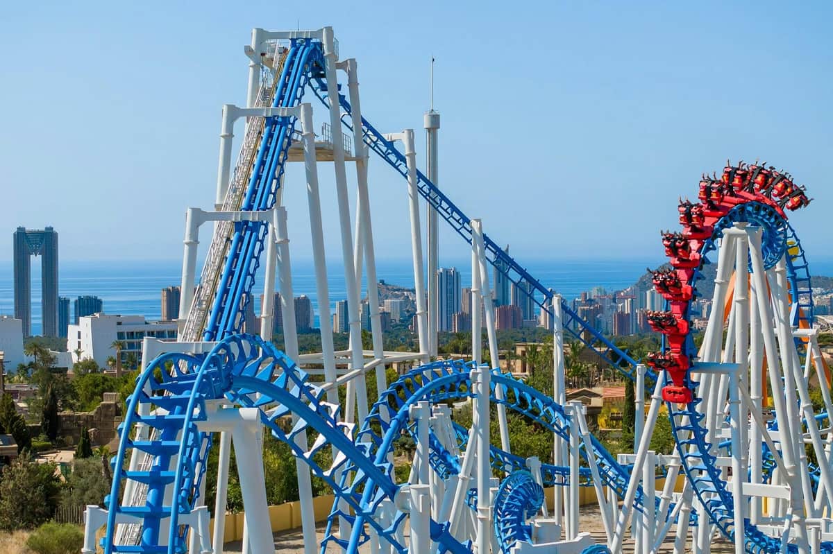 A blue and white rollercoaster with loops and drops, with a cityscape and ocean in the background under a clear sky.