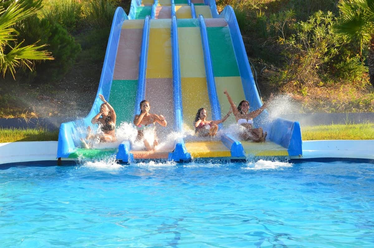 Four people splashing into a pool from parallel water slides of various colors on a sunny day.