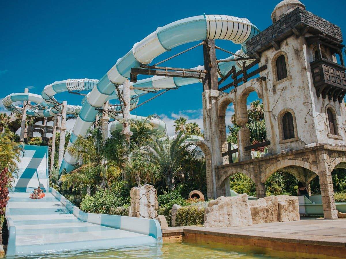 Outdoor water park with large blue and white tube slides, surrounded by palm trees and a stone structure, under a clear blue sky.
