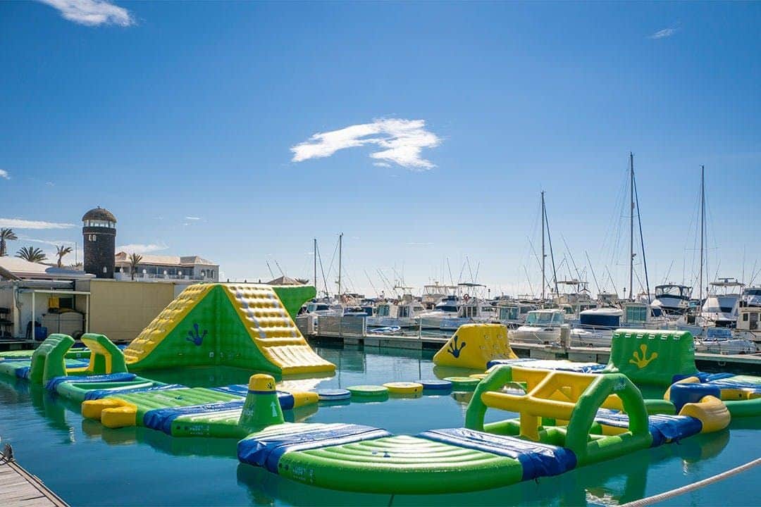 Inflatable water park with slides and obstacles floats on water, with a marina full of boats in the background under a clear sky.